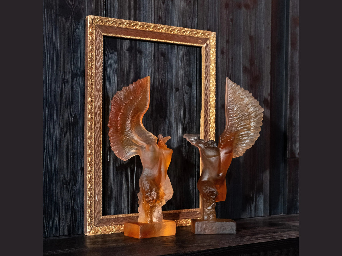 Two Daum Céyx glass sculptures of winged figures in front of an ornate gold frame.