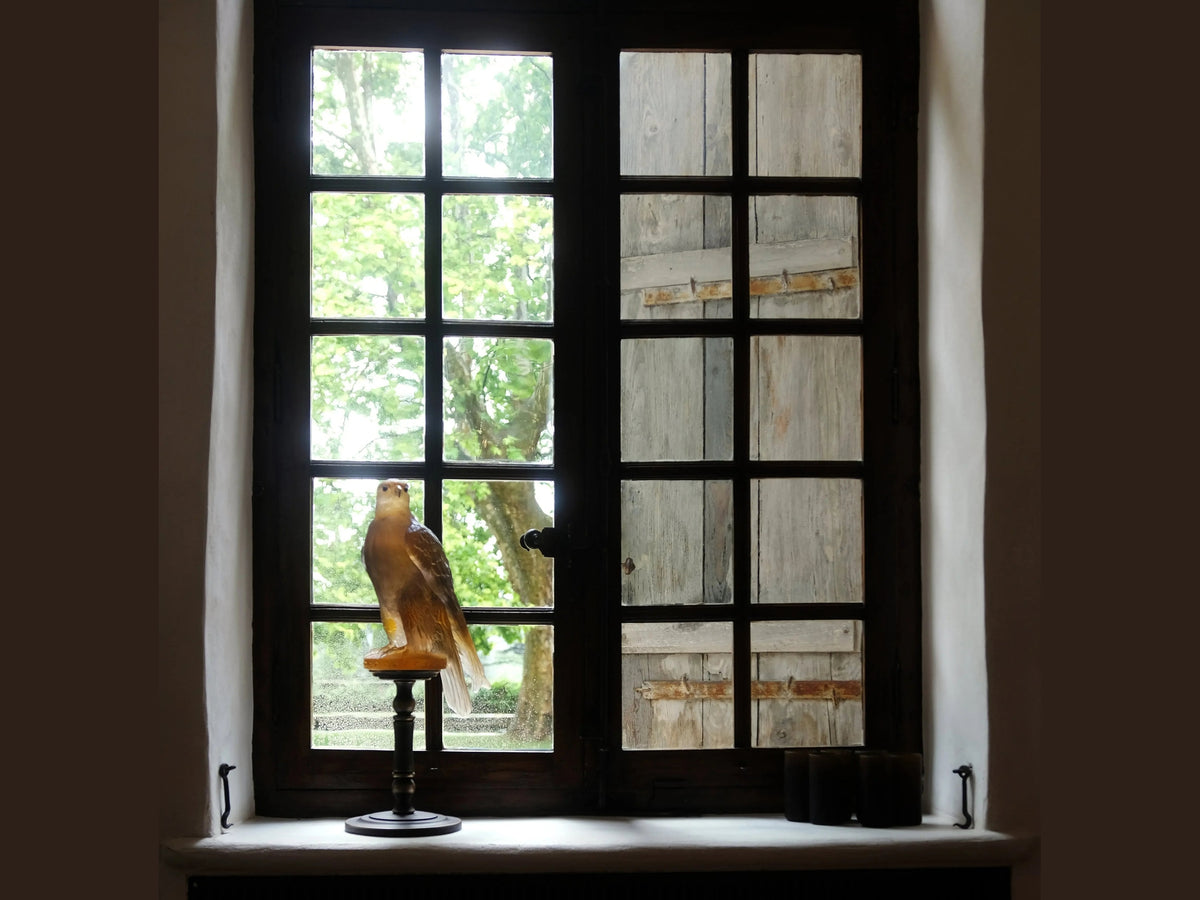 Daum Faucon Gerfaut Ambre-rose glass falcon sculpture on a stand by a window.