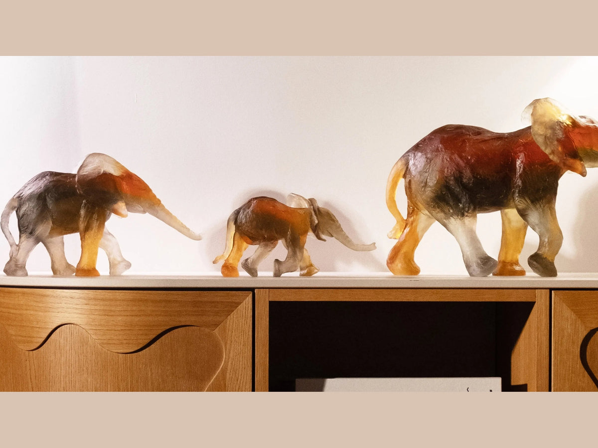 Three Daum Savannah Elephant glass sculptures in varying sizes on a wooden shelf.